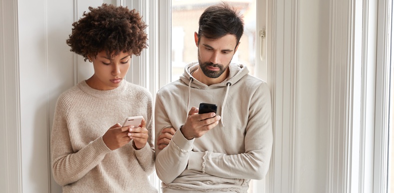 Horizontal shot of addicted sister and brother use wireless internet at home apartment, stand near window, enjoy day light, chat online on mobile phones, update profile in app. Technology concept